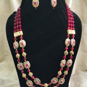 May include: A three-strand necklace and matching earrings set. The necklace features strands of small, round, red beads interspersed with ornate gold-colored oval beads with red center stones. The earrings have a similar design.