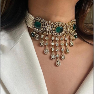 Könnte beinhalten: Ein goldfarbenes Choker-Halsband mit großen smaragdgrünen Edelsteinen und kleineren klaren Steinen. Die Halskette hat eine Perlenkette und baumelnde tropfenförmige Anhänger. Das Design ist aufwendig und elegant, passend für formelle Anlässe.
