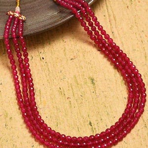May include: A three-strand necklace featuring small, faceted, ruby-red beads. The necklace is displayed on a neutral surface, showcasing the vibrant color and delicate design. The necklace is a jewelry item.