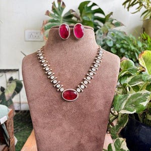 Puede incluir: Un conjunto de collar y pendientes exhibido sobre un busto marrón. El collar presenta una gran gema roja ovalada en el centro, rodeada de piedras claras más pequeñas. Los pendientes a juego tienen gemas rojas ovaladas. La joyería se presenta sobre un fondo de plantas verdes.