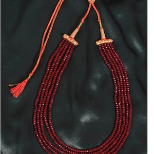 Puede incluir: Un collar de múltiples hilos con cuentas facetadas de color rojo intenso. El collar tiene un cordón naranja con una borla y detalles decorativos dorados. Las cuentas están dispuestas en múltiples hilos, creando un efecto de capas.