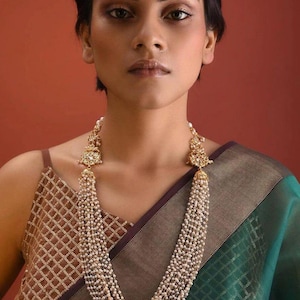 May include: A multi-strand pearl necklace with gold-toned accents. The necklace features a detailed pendant at the top and cascades down the chest. The model is wearing a brown and green patterned top.