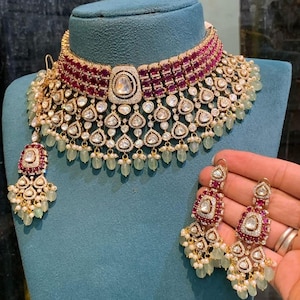 Könnte beinhalten: Ein goldfarbenes Halsketten- und Ohrring-Set. Die Halskette hat ein weinrotes Band, einen großen Mittelstein und tropfenförmige Akzente. Die Ohrringe spiegeln das Design der Halskette wider, mit weinroten und klaren Steinen und hängenden grünen Perlen. Der Schmuck wird auf einer blauen Büste präsentiert.