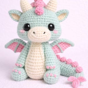 Könnte beinhalten: Ein gehäkeltes Drachenplüschtier in Pastellfarben. Der Drache hat einen mintgrünen Körper, einen cremefarbenen Bauch und rosa Akzente auf den Flügeln, Hörnern und dem Schwanz. Er hat große schwarze Augen und einen freundlichen Ausdruck. Der Drache ist etwa 20 cm groß.