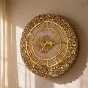 Puede incluir: Un reloj de pared redondo con marco dorado y números romanos. La esfera del reloj es de color beige claro con detalles dorados. El marco exterior está incrustado con pequeñas piedras doradas brillantes. Las manecillas del reloj son doradas.