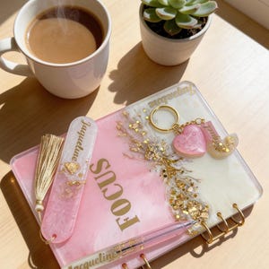 May include: A pink and white resin journal with the word "FOCUS" in gold lettering. A matching pen, bookmark, and keychain with gold accents and the name "Jacqueline" are also present. A cup of coffee and a succulent plant are in the background.