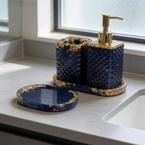 Könnte beinhalten: Ein marineblaues Badezimmer-Set mit goldenen Akzenten. Das Set umfasst einen Seifenspender, einen Zahnbürstenhalter und eine kleine Ablage. Die Artikel haben ein geometrisches Muster und Goldblattdetails.