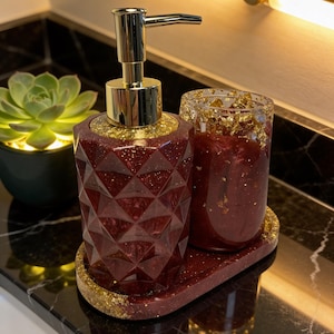 Può includere: Un set da bagno decorativo con un dispenser di sapone, un bicchiere e un vassoio bordeaux. Il set presenta un design geometrico con dettagli dorati. Il dispenser di sapone ha una pompa dorata. Il bicchiere e il vassoio sono anch'essi bordeaux con scaglie d'oro.