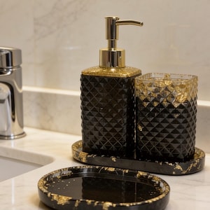 May include: A bathroom accessory set featuring a black soap dispenser with a gold pump, a matching tumbler, and an oval soap dish. Each piece has a textured geometric diamond pattern and is accented with gold leaf flakes throughout the design.