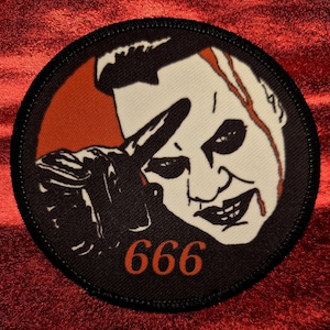 May include: Round patch with a black border featuring a stylized illustration of a figure with white skin, black eyes, and red accents. The figure is pointing with a gloved hand. The number "666" is printed in orange.