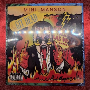 Puede incluir: Portada del álbum "Git Dead" de Mini Manson. La ilustración muestra una figura de dibujos animados con gafas de sol y un traje, mostrando el dedo medio. El fondo incluye llamas, rayos y las palabras "Las Vegas".