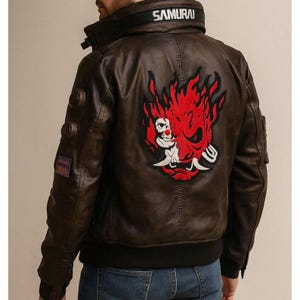May include: A brown leather jacket with a black collar and cuffs. The back features a large red and white graphic design. The word "SAMURAI" is printed on the collar. The jacket has multiple pockets and patches.