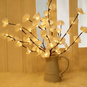 Puede incluir: Un arreglo floral decorativo con flores blancas iluminadas sobre tallos marrones, dispuesto en un jarrón gris claro. El jarrón tiene un asa y la palabra "FLOWERS" está impresa en la parte delantera. El arreglo está sobre un fondo suave y cálido.