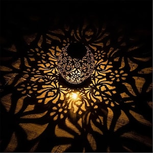 Puede incluir: Una linterna decorativa de metal con intrincados recortes florales, que proyecta una luz cálida y estampada sobre una superficie de madera. El diseño de la linterna crea un juego de luces y sombras, realzando el ambiente.