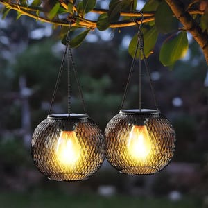 May include: Two hanging solar-powered lanterns with a black metal mesh design. Each lantern has a warm, glowing lightbulb inside, suspended from a chain and hook. The lanterns are hanging from a tree branch with green leaves, creating a cozy outdoor ambiance.