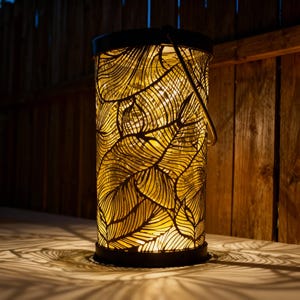 Puede incluir: Una linterna cilíndrica con una estructura de metal negro y un diseño de hojas. La linterna tiene un asa y emite un brillo cálido y dorado, creando sombras con forma de hojas. La linterna mide unos 30 cm de altura.