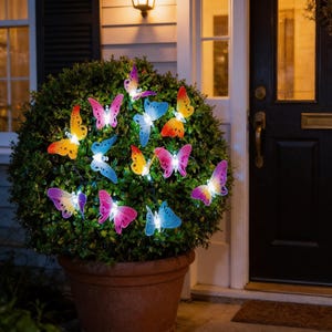 May include: A potted green bush adorned with colorful, illuminated butterfly lights. The butterflies are various shades of pink, blue, yellow, and orange, creating a whimsical and decorative display. The lights are strung throughout the bush, adding a warm glow.