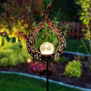 May include: A solar-powered garden light with a decorative design. The teardrop-shaped metal frame has a dark bronze finish with cut-out patterns. A crackled glass orb glows within the frame. The light is mounted on a black stake.