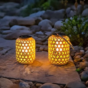 May include: Two illuminated bamboo lanterns with a geometric pattern. The lanterns have a warm, yellow glow and are sitting on a stone surface. The lanterns have a black top and a woven handle.
