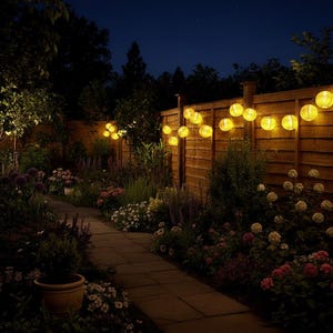 Puede incluir: Una escena de jardín nocturna con un camino de piedra que atraviesa parterres de flores. Faroles de papel amarillos están colgados a lo largo de una valla de madera, iluminando el jardín. El cielo oscuro sirve de telón de fondo a las luces brillantes y la exuberante vegetación.