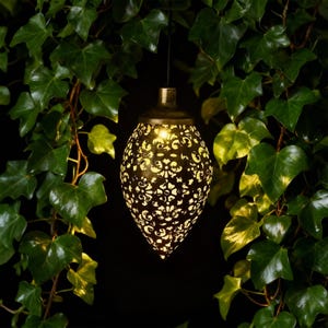 May include: A decorative hanging lantern with an intricate, openwork pattern. The lantern is a warm gold colour and is suspended by a thin black wire. The background is a dark green with leafy vines.