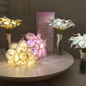May include: Several bouquets of artificial calla lilies with integrated LED lights, featuring white, yellow, and pink petals. The flowers are arranged in clear glass vases tied with ribbons, and a rectangular box labeled Calla Bouquet LED lights.
