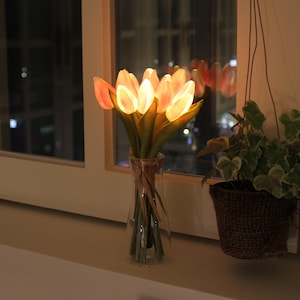 May include: A glass vase holds a bouquet of illuminated artificial tulips. The tulip petals glow with a soft orange light, contrasting with the green stems. The vase is tied with twine and sits on a windowsill near a hanging potted plant.