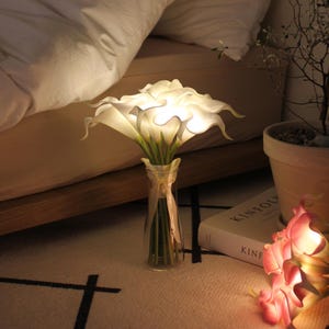 May include: A bouquet of white calla lilies in a clear glass vase tied with a ribbon. The flowers are illuminated, creating a soft glow. The vase sits on a patterned rug near a bed.