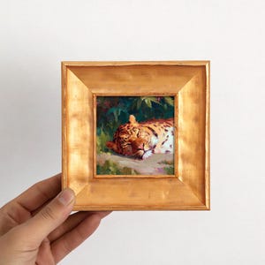 May include: A framed painting of a resting leopard. The artwork features a detailed depiction of the animal with orange and black spots, set against a backdrop of green foliage. The frame is a square shape with a gold finish.