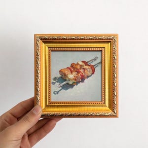 May include: A small, square, framed painting featuring two skewers of grilled food. The painting is set in a gold-colored frame. The skewers have pieces of meat and vegetables. The painting is held by a hand against a white background.