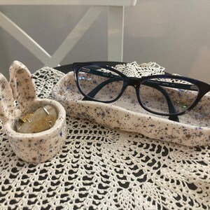 Può includere: Un portagioie e un vassoio per occhiali in ceramica maculata. Il portagioie ha la forma di una mano con tre dita e un pollice. Il vassoio è rettangolare. Entrambi sono bianco sporco con macchie blu. Un paio di occhiali con montatura nera poggia sul vassoio.