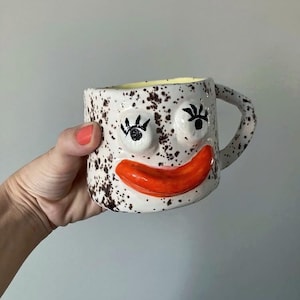 Może przedstawiać: Kapryśny ceramiczny kubek z uśmiechniętą twarzą. Kubek ma białą podstawę w brązowe plamki, jasnopomarańczowe usta i czarne rzęsy. Wnętrze jest żółte, a uchwyt ma fakturę.