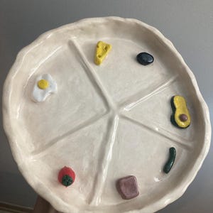 Könnte beinhalten: Ein handgefertigter, cremefarbener Keramikteller mit sechs Fächern, die jeweils ein Miniatur-Lebensmittel enthalten. Die Lebensmittel umfassen ein Spiegelei, Käse, eine Olive, Avocado, eine Gurke und Toast. Der Teller ist rund.