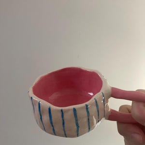 Peut inclure: Une tasse en céramique faite à la main avec un intérieur et des anses roses. L'extérieur est blanc avec des rayures bleues verticales. La tasse est tenue par une main. La tasse est une œuvre d'art unique.