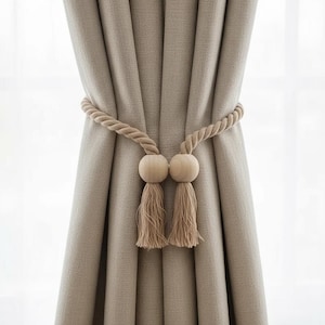 May include: Neutral-toned linen curtains held by a rope tieback. The tieback features wooden ball details and matching tassels. The thick, twisted rope adds texture. The curtains create a soft, natural look, suitable for various interior styles.