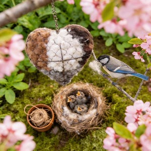 May include: A heart-shaped bird feeder with brown, white, and gray wool filling, suspended by a chain. A small bird perches nearby, gathering fluff. A nest with baby birds and a small bowl of twine are also visible.