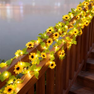 Puede incluir: Una barandilla de madera adornada con una guirnalda de girasoles amarillos y hojas verdes. Pequeñas luces blancas cálidas se entrelazan con las flores, creando un brillo suave. La barandilla es de madera marrón, con un fondo borroso de agua.