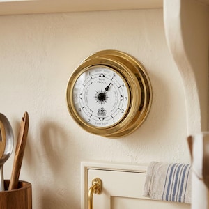 May include: A circular tide clock with a polished brass frame and a white face. The dial features black markings, a central compass rose, a small ship illustration, and text reading TIDE CLOCK, HIGH TIDE, LOW TIDE, RISING TIDE, and FALLING TIDE.