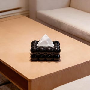Puede incluir: Un dispensador de pañuelos negro con un diseño de burbujas, que contiene un pañuelo blanco. El dispensador está colocado sobre una mesa de madera marrón claro. El diseño del dispensador está hecho de esferas negras brillantes.
