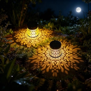 Puede incluir: Dos luces solares cilíndricas para jardín con intrincados recortes florales proyectan patrones en el suelo. Las luces están encendidas, creando un brillo cálido en un jardín. La escena está ambientada de noche con una luna llena.