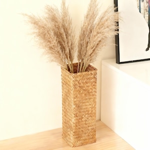 Puede incluir: Un jarrón alto y rectangular de mimbre tejido lleno de hierba de la pampa beige seca y esponjosa. El jarrón está hecho de material tejido de color natural y se encuentra sobre una superficie de madera de color claro. Es un artículo de decoración del hogar.