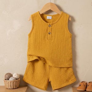 May include: Mustard yellow two-piece outfit for a child, consisting of a sleeveless top with two buttons and matching shorts. The top has the word "TROOH" printed on the label. The outfit is made of a textured fabric and is hanging on a wooden hanger.