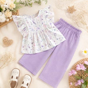 Può includere: Un completo da due pezzi per bambini, composto da un top bianco con stampa floreale e maniche a balze e pantaloni color lavanda. L'outfit è abbinato a sandali bianchi. Il top ha uno scollo tondo e i pantaloni hanno una vita elasticizzata.