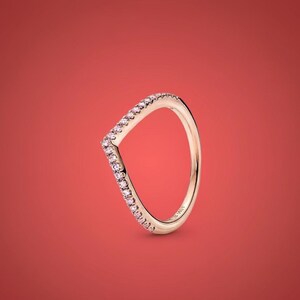 May include: A gold-colored ring with a V-shaped design. The ring is adorned with small, pink gemstones along the V-shape. The ring is set against a solid red background.