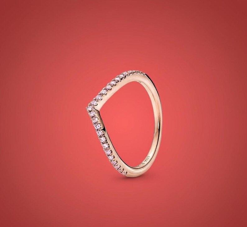 May include: A gold-colored ring with a V-shaped design. The ring is adorned with small, pink gemstones along the V-shape. The ring is set against a solid red background.