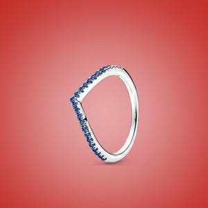 May include: A silver-colored ring with a V-shaped design. The ring is adorned with small, round, blue gemstones set along the V-shape. The ring is set against a red background.