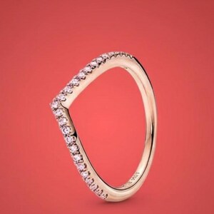 May include: A rose gold V-shaped ring with a row of small pink gemstones along the top edge. The ring is set against a solid red background, highlighting its elegant design. The ring is a piece of jewelry.