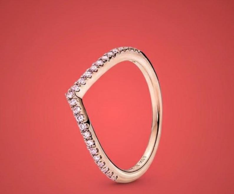 May include: A rose gold V-shaped ring with a row of small pink gemstones along the top edge. The ring is set against a solid red background, highlighting its elegant design. The ring is a piece of jewelry.