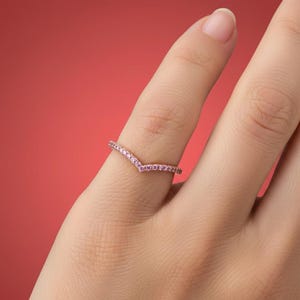 May include: A delicate, V-shaped ring with small, pink gemstones set along the band. The ring is displayed on a finger against a solid red background. The ring's design is simple and elegant, suitable for everyday wear or special occasions.