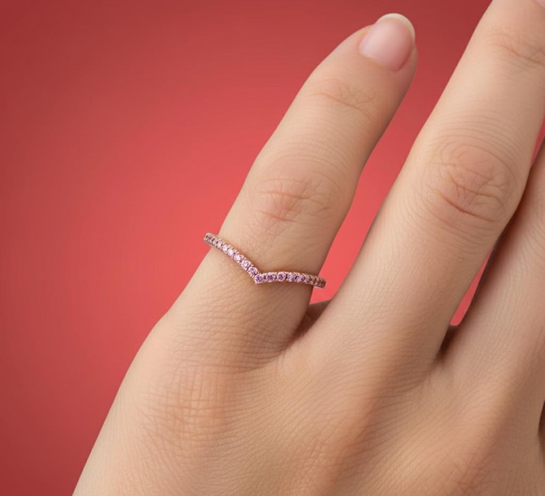May include: A delicate, V-shaped ring with small, pink gemstones set along the band. The ring is displayed on a finger against a solid red background. The ring's design is simple and elegant, suitable for everyday wear or special occasions.